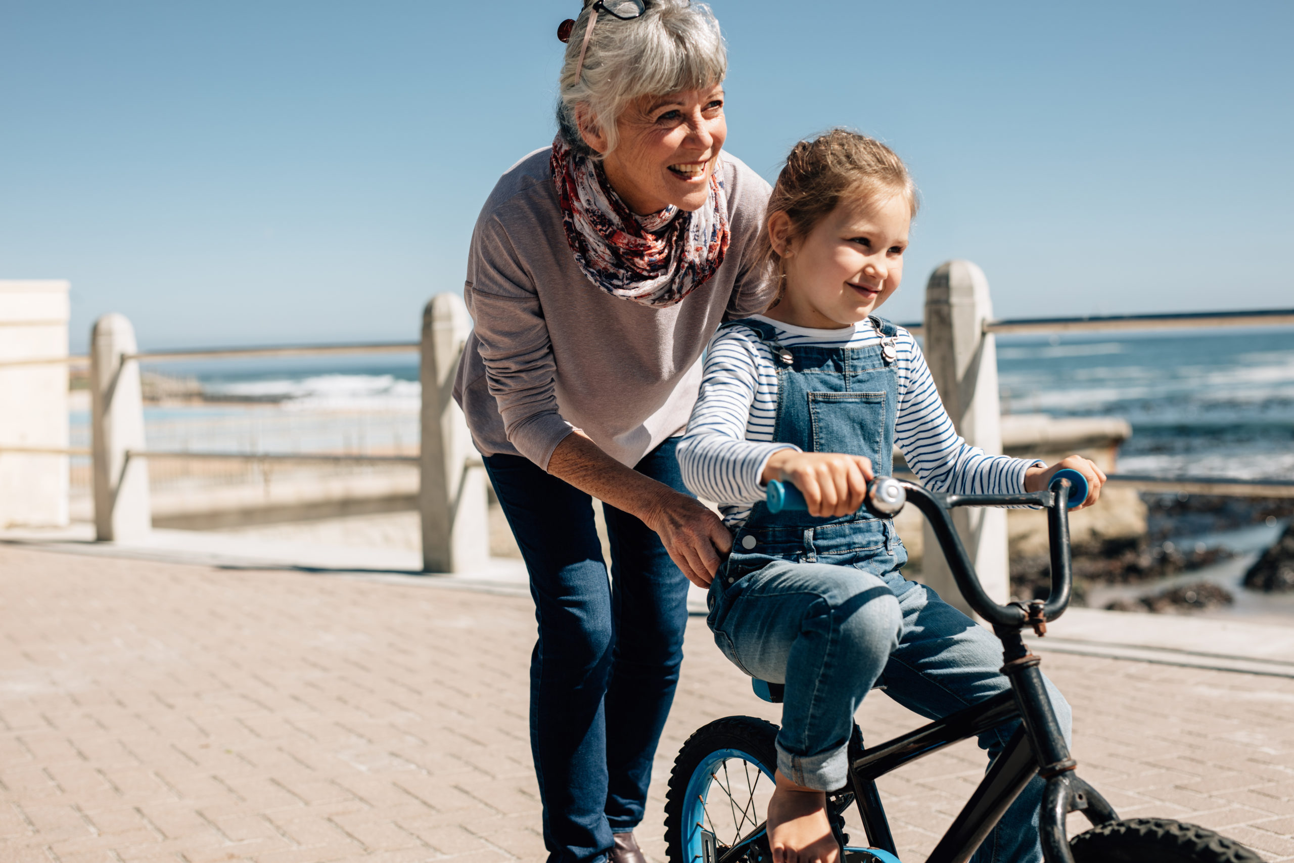 Girl trying to ride a bicycle with the help of her grandmother at seaside promenade. Senior woman holding the bicycle while her granddaughter learns to ride it.