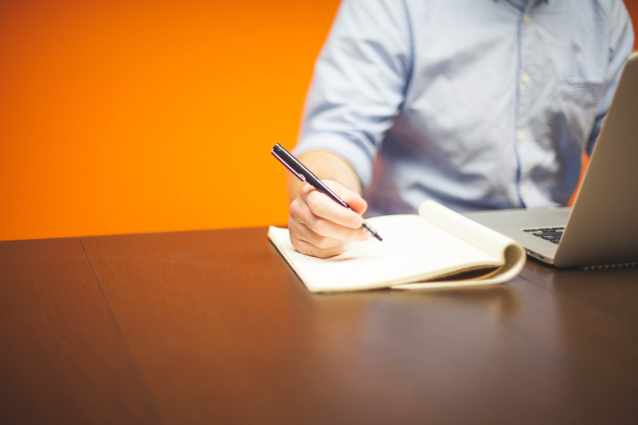 man-sitting-at-table-working-on-laptop-and-writing-in-notebook