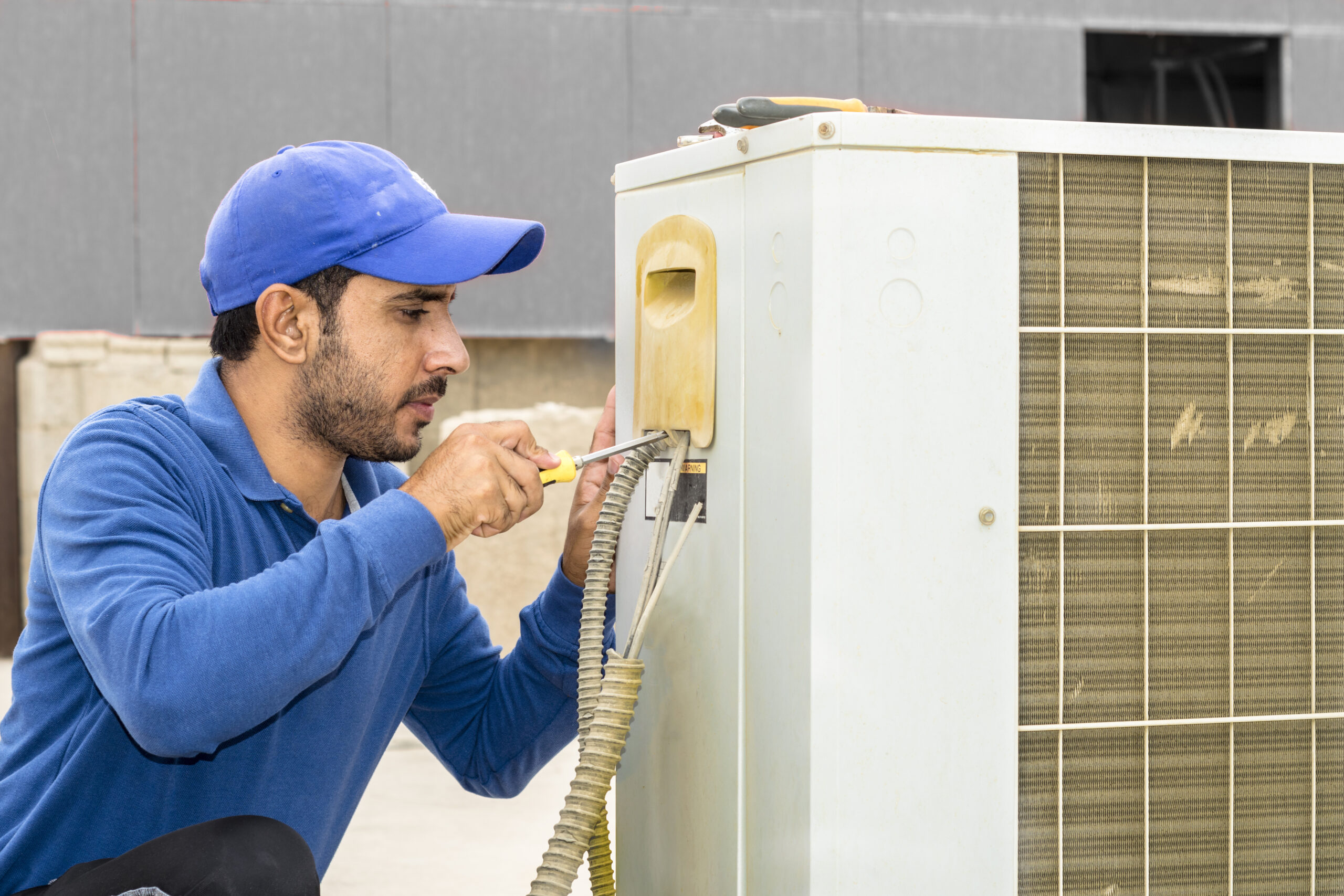 a professional electrician man is fixing a heavy duty unit of central air conditioning system by his tools on the roof top and wearing grey color of uniform and white cap a professional electrician man is fixing a heavy duty unit of central air conditioning system by his tools on the roof top and wearing grey color of uniform and white cap